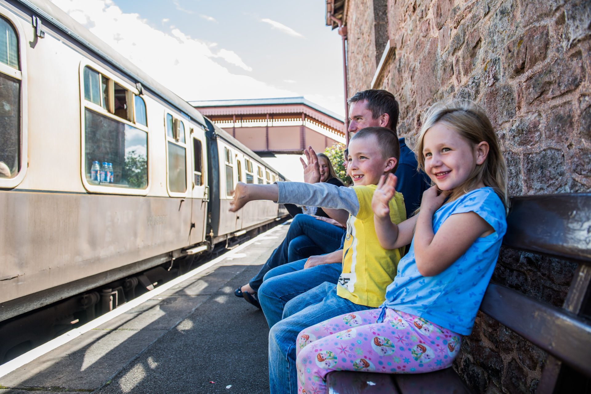 A young family sit on a bench on a platform and wave at a train